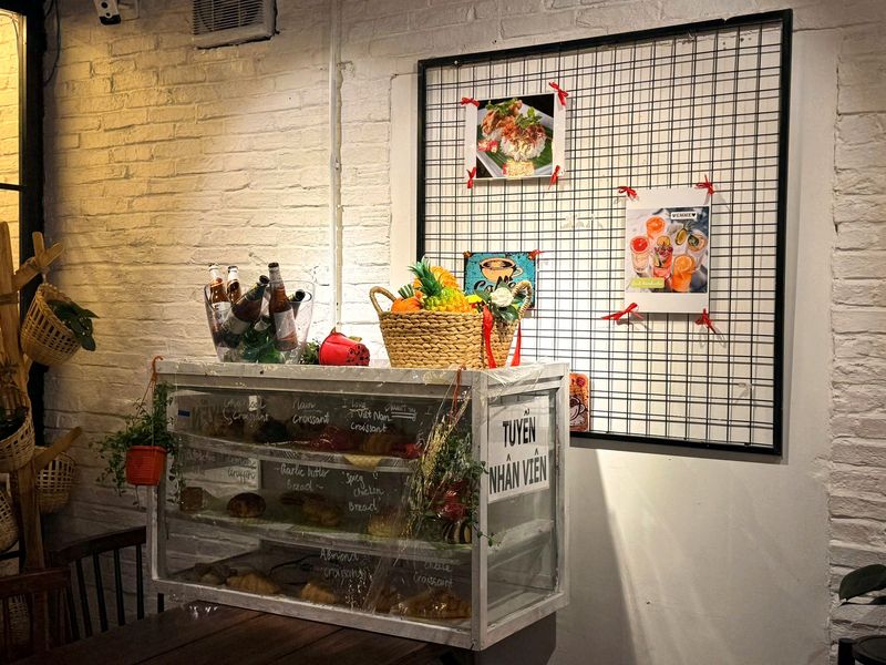 Tủ kính bày bánh đặt cạnh khung lưới treo tranh món ăn, bên cạnh dán tờ tuyển nhân viên
