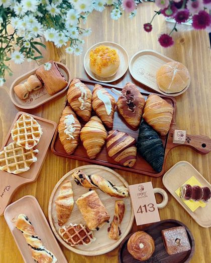 Nhìn từ trên xuống mẻ bánh trong ngày: croissant, bánh cuộn, waffle và bánh mì trên những khay gỗ khắc EMME, quanh khối gỗ số 413