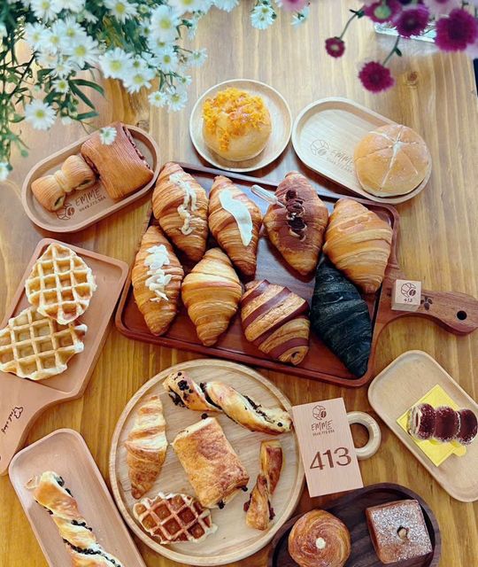 Nhìn từ trên xuống mẻ bánh trong ngày: croissant, bánh cuộn, waffle và bánh mì trên những khay gỗ khắc EMME, quanh khối gỗ số 413