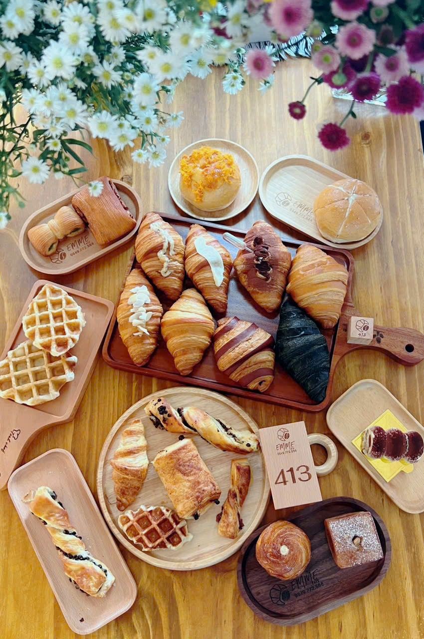 Nhìn từ trên xuống mẻ bánh trong ngày: croissant, bánh cuộn, waffle và bánh mì trên những khay gỗ khắc EMME, quanh khối gỗ số 413
