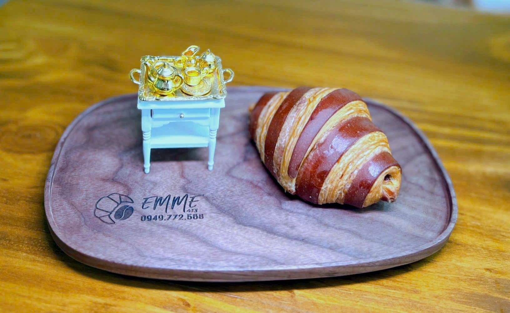Croissant sọc socola trên đĩa gỗ khắc chữ EMME 413, bên cạnh là bộ bàn trà tí hon mạ vàng