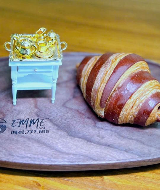 Croissant sọc socola trên đĩa gỗ khắc chữ EMME 413, bên cạnh là bộ bàn trà tí hon mạ vàng