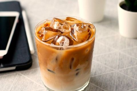 Ly cà phê sữa đá trong ly thuỷ tinh thường, cà phê loang thành vân nâu quanh đá, bên cạnh là cuốn sổ và điện thoại