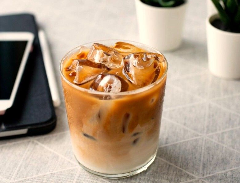 Ly cà phê sữa đá trong ly thuỷ tinh thường, cà phê loang thành vân nâu quanh đá, bên cạnh là cuốn sổ và điện thoại
