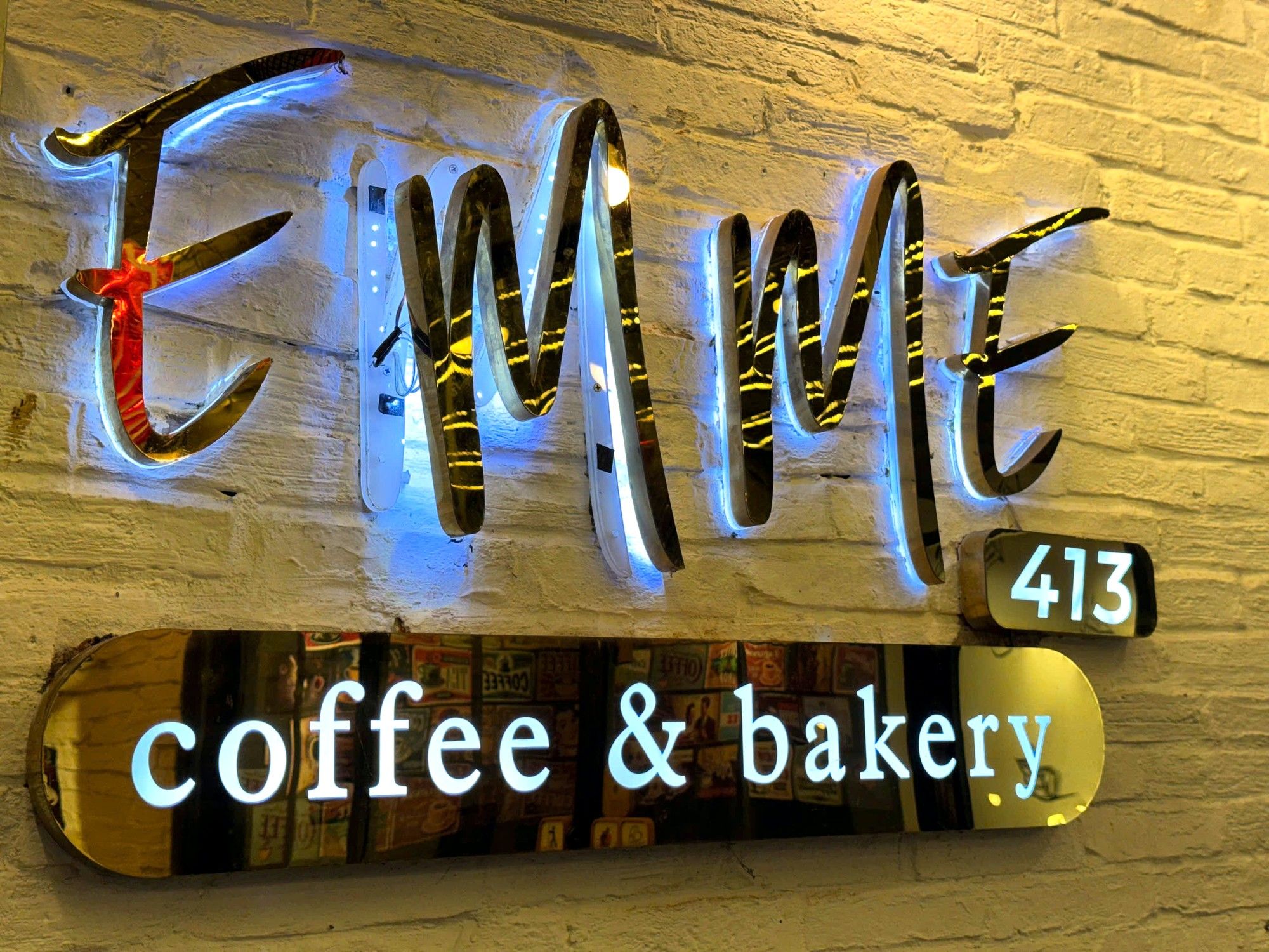 Bảng hiệu EMME 413 coffee & bakery gắn trên tường gạch, chữ viền đèn xanh